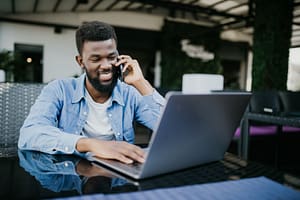 young Black man talking on cell phone while sitting at a cafe with a laptop