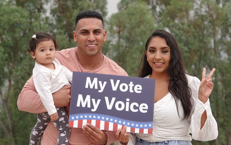 Woman with man holding a toddler. They are holding a sign: My Vote My Voice