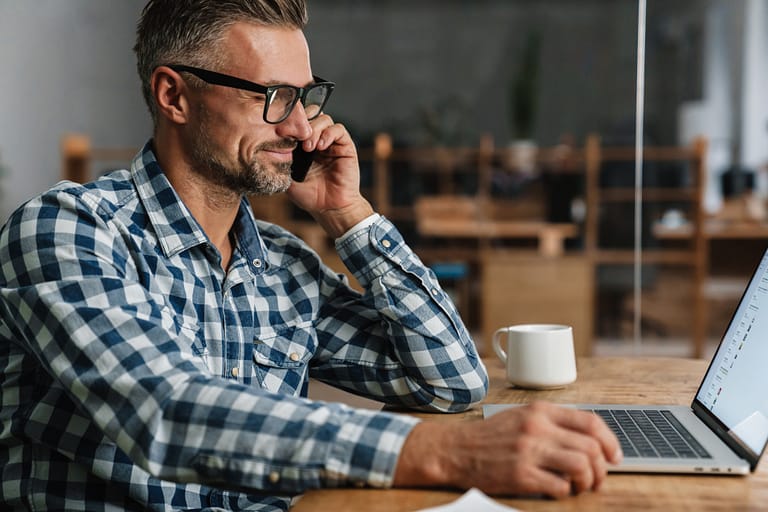 Smiling grey haired man talking on mobile phone while working with laptop