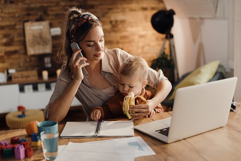 Working mother feeding her son with a banana while talking on the phone at home.