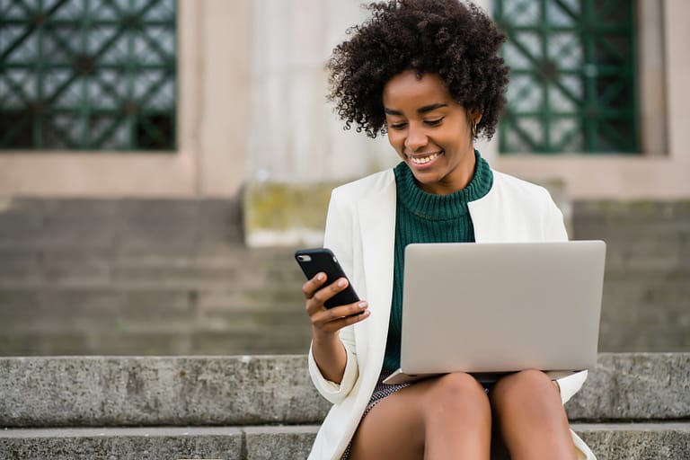 Business woman using her mobile phone and laptop outdoors.