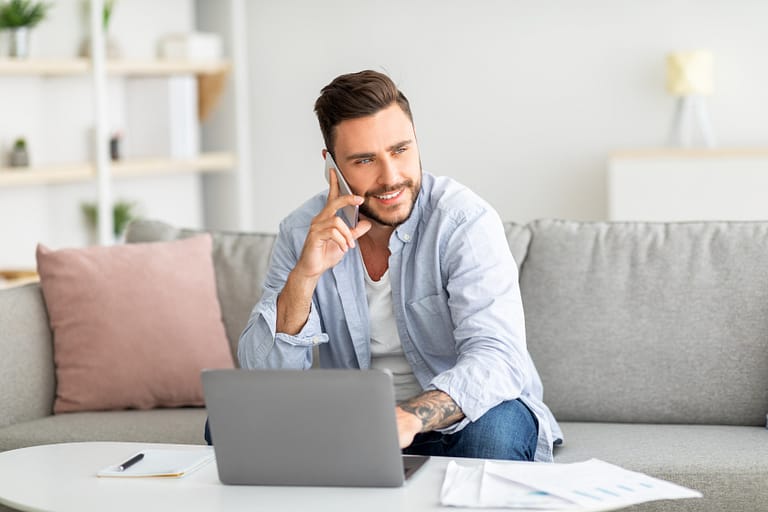 light skinned man talking on cellphone and working on laptop from home, sitting on sofa