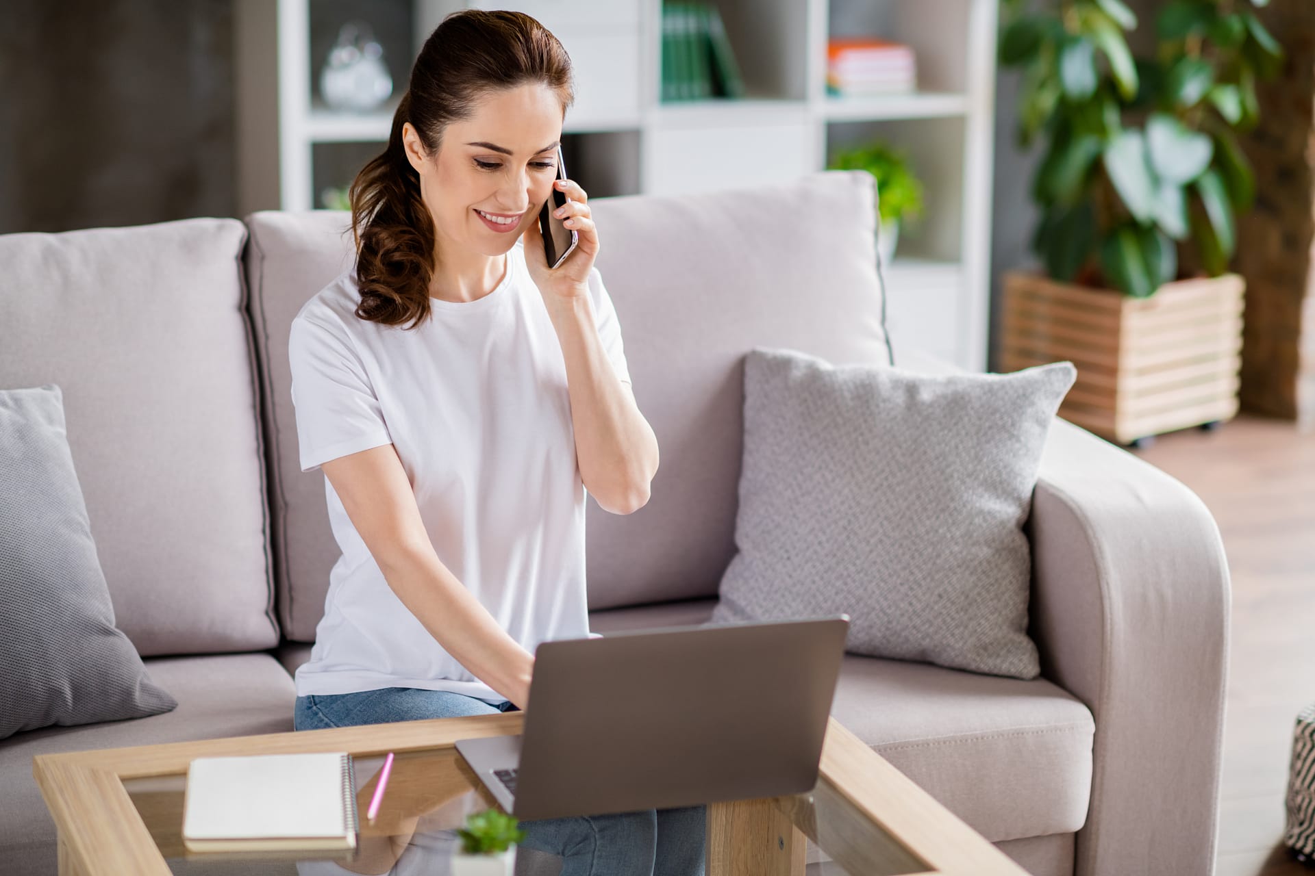 young woman sitting on sofa speak phone use laptop working from home indoors